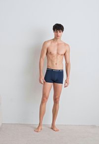 Mørkeblå boxershorts med merkebånd, laget av et glatt, elastisk materiale, iført av en stående mannlig modell på et lyst gulv.