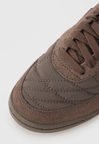 Pantofii sport maro au un exterior din suede texturat și nylon matlasat, cu șireturi plate și talpă din cauciuc cu detalii cusute.