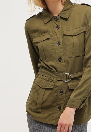 Frau trägt eine olivgrüne Utility-Jacke mit Gürtel, Klappentaschen und schwarzen Knöpfen, kombiniert mit grauen Strickhosen.