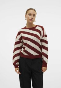 Maglione a righe in rosso e bianco sporco, realizzato in tessuto morbido, con collo rotondo e spalle scese. Abbinato a pantaloni neri.