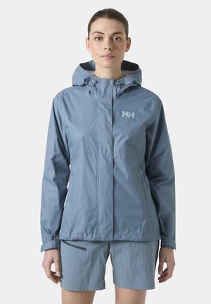 Mujer con chaqueta impermeable con capucha de color azul claro y pantalones cortos a juego, de pie contra un fondo claro y liso.