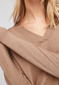 Jasno-brązowy sweter w serek o miękkiej fakturze z prążkowanym wykończeniem przy dekolcie i mankietach, z luźnymi rękawami.