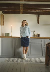 Maglione azzurro chiaro a maglia con colletto rotondo e bottoni, abbinato a una gonna di jeans e calzini bianchi, ambientato in una cucina con dettagli in legno.