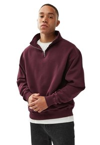 Burgundin värinen fleece-huppari, jossa on neljännesvetoketju, resorit hihoissa ja helmassa. Päällä valkoinen paita, jossa rento istuvuus ja pehmeä rakenne.