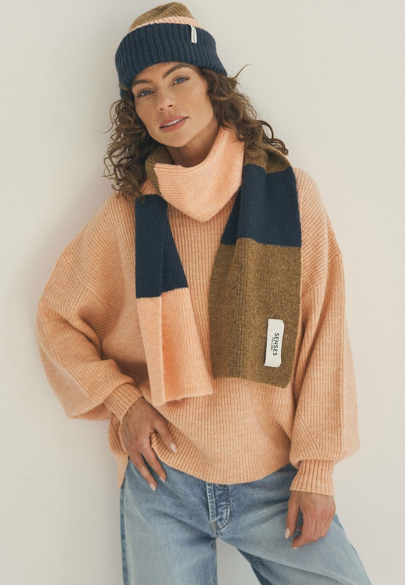Soft pink oversized sweater with ribbed texture, paired with a multicolored scarf featuring navy and brown stripes. Complete with a matching beanie.