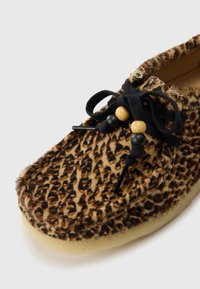 Sleveland genser i leopardmønster laget av myk pels, med avrundet form, svarte snører og dekorative treperler. Lys farge på sålen.