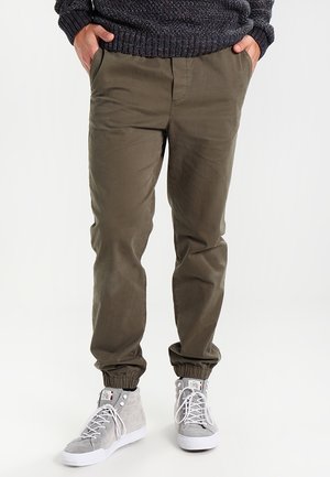 Mies, jolla on päällään oliivinvihreät vapaa-ajan housut joustavilta resoreilta, harmaat korkeavartiset tennarit ja tumma teksturoiduista kankaista valmistettu neule, kädet taskuissa.