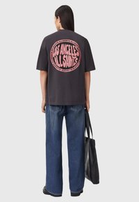 Czarny t-shirt z krótkim rękawem, z dużą okrągłą różową grafiką na plecach z napisem "Los Angeles AllSaints". Noszony z niebieskimi spodniami w luźnym stylu.