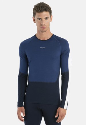 Junger Mann trägt ein eng anliegendes langärmliges Sportshirt in Marineblau mit einem weißen Streifen auf dem rechten Arm und steht vor einem einfarbigen Hintergrund.