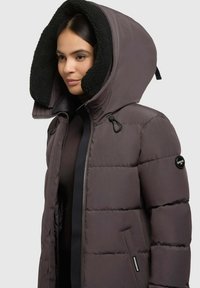Chaqueta de plumón marrón para mujer con capucha forrada de felpa negra, cremallera frontal y logo de parche en la manga. Diseño texturizado e insulated.