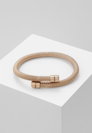 Pulsera de alambre flexible en tono dorado con dos extremos cilíndricos, con marca grabada. Textura suave con diseño en espiral.