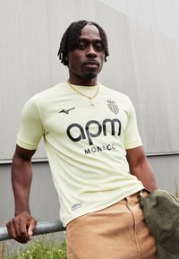 Muž s krátkymi dredmi sa opiera o zábradlie, má na sebe svetložltý dres AS Monaco a drží zelenú čiapku.