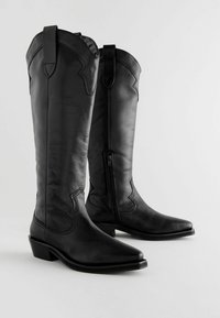 Bottes de cowboy en cuir noir avec un bout pointu, des détails de couture courbés et une fermeture éclair sur le côté. Elles possèdent un petit talon en bloc et des tirants.