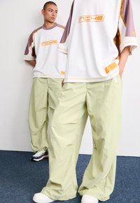 Twee modellen dragen oversized witte en paarse T-shirts met korte mouwen met oranje accenten, gecombineerd met lichtgroene losse broeken en sneakers.