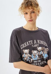 Grijze katoenen T-shirt met een graphic van vier beren in blauwe outfits, met de tekst "CREËER EEN VRIENDELIJKERE PLANEET" in een opvallende, lichte kleur.