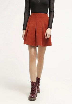 Czerwona zamszowa spódnica rozszerzana z plisami, zestawiona z czarną przezroczystą bluzką z długim rękawem oraz bordowymi sznurowanymi botkami do kostki. Gładka struktura, dopasowana talia.