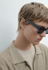 Schwarze rechteckige Sonnenbrillen mit dicken Rahmen, kombiniert mit einem beigen strukturierten Hemd mit Kragen und einem leichten Pullover. Kurze, zerzauste Haare sind sichtbar.