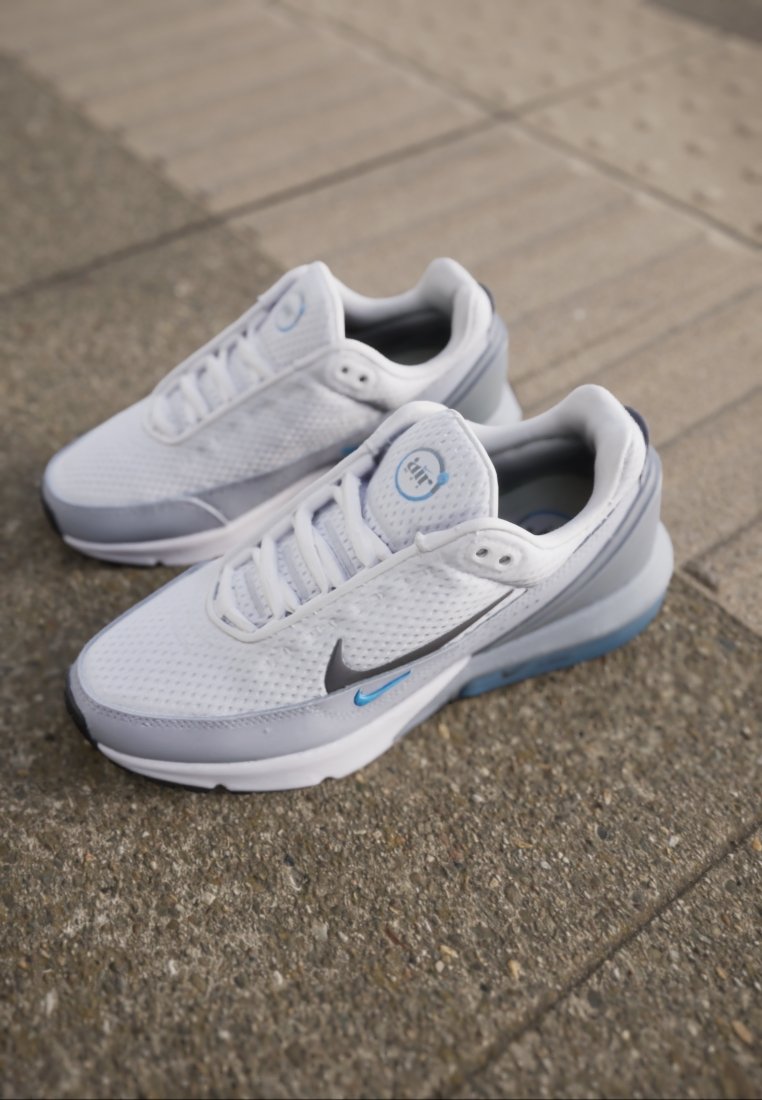 Baskets Nike Air Max blanches et grises avec des accents bleus sur un pavé texturé et des dalles de guidage.