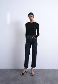 Femme debout contre un mur uni, portant un haut à manches longues noir, un jean large bleu foncé et des sandales à talons noires.