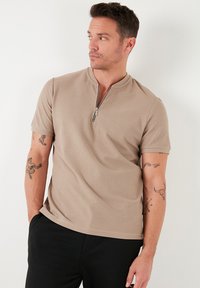 Beige, cipzárral ellátott, rövid ujjú pólóing, texturált anyagból készült. Félcipzáros fazonú, laza illeszkedésű, sötét nadrággal párosítva.