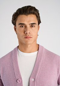 Vaaleanpunainen neulottu neuletakki, jossa on resorillinen huivikaulus, edessä napitus ja päällä valkoinen pyöreäkauluksinen t-paita. Sileä pinta, ilman kuvioita.