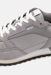 Baskets grises en daim et tissu avec lacets blancs et logo "BOSS" noir sur le côté et la languette, vues de côté en gros plan.