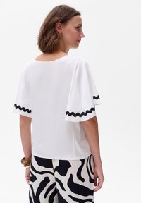 Biała bluzka z krótkim rękawem, z czarną falistą lamówką przy rękawach, gładki materiał, okrągły dekolt, zestawiona z czarno-białymi spodniami w wzory.