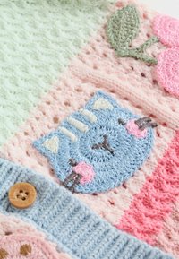 Megzta tekstilė su pastelinėmis žalia, rožine ir mėlyna dalimis, mėlynu katino motyvu ir mediniu mygtuku. Minkšta, tekstūruota medžiaga su išsiuvinėjimu.