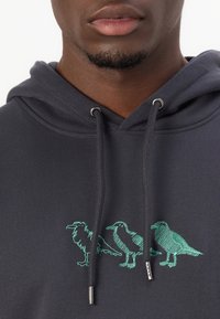 Kapuzenpullover aus strukturiertem Material in Anthrazit, mit einem tealfarbenen gestickten Design von drei Vögeln auf der Vorderseite und silbernen Akzenten an der Kordel.