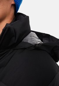 Sort isoleret jakke med lynlås-kant, der har et gråt foer og justerbar snor ved halsen. Glat ydertekstur.