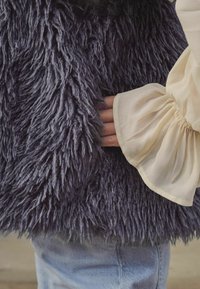 Pelēks, shaggy mākslīgā pūka veste ar garu, mīkstu tekstūru, valkāta virs krēmkrāsas blūzes ar pūkainām piedurknēm un savelkamiem aprocēm.