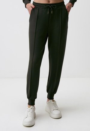 Persoon draagt zwarte taps toelopende joggebroek met elastische boorden en witte sneakers, handen in de zakken, staand op een effen witte vloer.