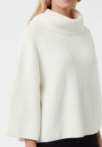 Kobieta nosząca luźny, biały sweter z prążkowanej dzianiny o szerokich rękawach i złożonym kołnierzem typu golf na jednolitym tle.