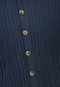 Merineel kootud kardigan, millel on vertikaalsed nöörmustrid ja viis ümmargust pruuni nuppu ees. Pehme tekstuur ja sooniline disain.