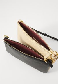 Due piccole pochette in pelle con zip, una beige e una nera, con interni color bordeaux e dettagli dorati, mostrate aperte e sovrapposte.