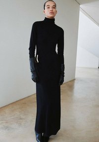 Robe longue noire à col roulé avec manches longues, silhouette ajustée et poche latérale. Associée à des gants en cuir noirs pour un contraste.