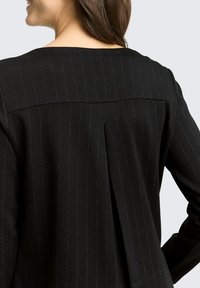 Czarna marynarka w prążki z plisą z tyłu i prostym dekoltem. Tkanina ma subtelny połysk i gładką fakturę.