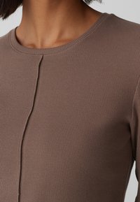 Närbild av en person som bär en brun ribbad stickad topp med rund halsringning och en vertikal sömdetalj ner genom mitten fram.