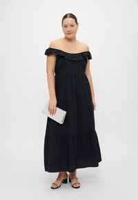 Juodas off-shoulder maksi suknelė su raukiniais ant kaklo, sluoksniuota sijoną ir iki kulkšnių siekiančiu modeliu. Modelis laiko sidabrinę rankinę ir vilki šlepetes su dirželiu.