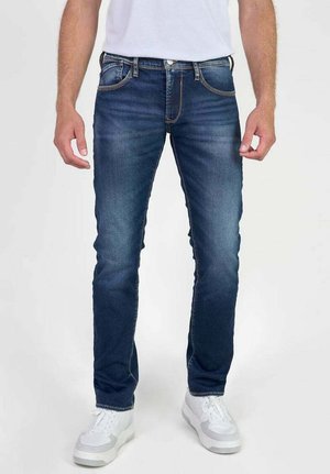 Jeans de hombre de denim azul oscuro con corte recto, ligero desvanecimiento, costuras naranjas y cinco bolsillos. Llevados con zapatillas blancas.