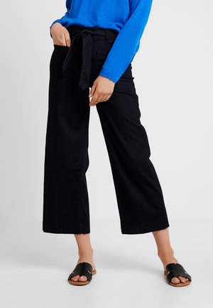 Nainen pukeutunut mustiin leveälahkeisiin lyhennettyihin housuihin, joissa on vyö, siniseen pitkähihaiseen yläosaan ja mustiin avojalkaisiin liukusandaaleihin.