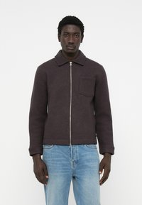 Dark gray wool jacket with a zip closure, oversized collar, and a chest pocket, worn over light blue denim jeans. Smooth texture.