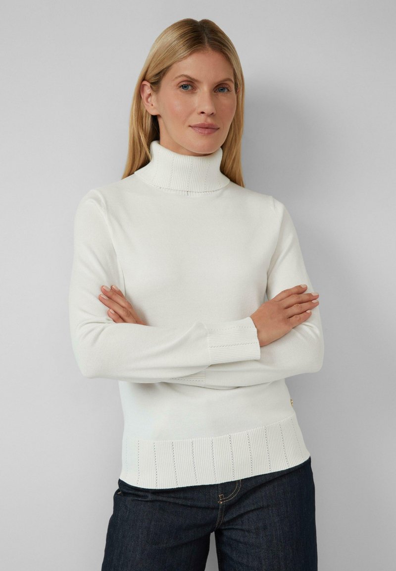 Pull en col roulé blanc avec une texture côtelée, présentant un design ajusté et des détails en tricot plat au niveau des poignets et de l'ourlet. Porté avec un jean en denim sombre.