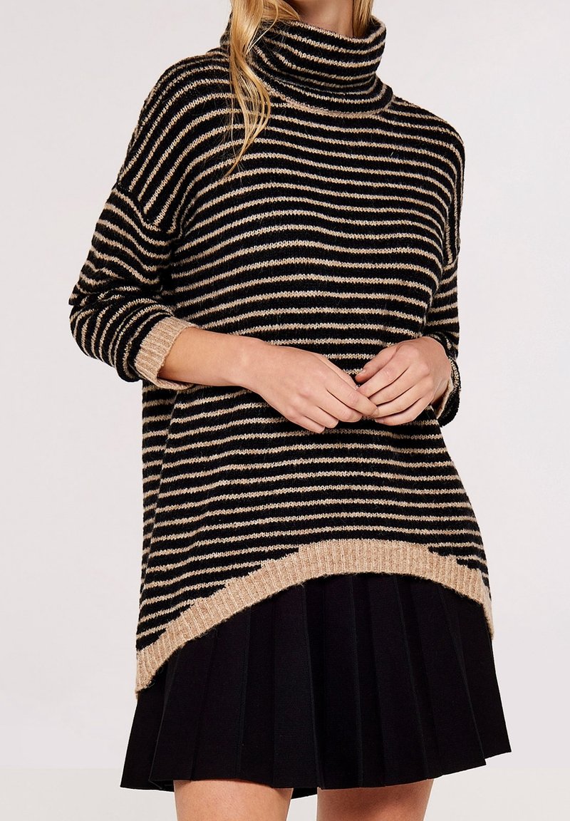 Femme portant un pull à col roulé rayé noir et beige avec une bordure beige et une jupe plissée noire, les mains légèrement jointes devant.