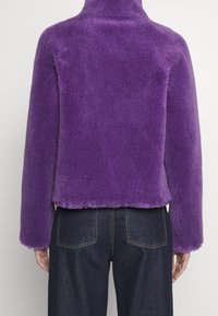 Lila fuskpälskappa med hög krage, som har en mjuk, strukturerad yta och en kort design. Matchas med mörka jeans.