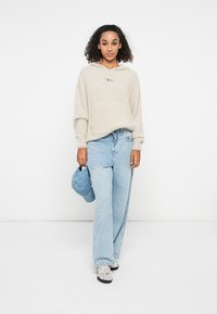 Beige, fluffy hoodie met een voorzak en geborduurd detail, gecombineerd met lichtblauwe wijde jeans. Houdt een blauwe pet vast.
