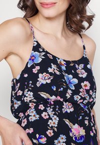 Femme portant une robe noire sans manches avec des motifs floraux bleus, roses et blancs et des fines bretelles.