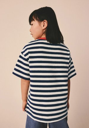 Dieťa s rovnými čiernymi vlasmi, oblečené v krátkomonkávovom tričku s námorníckymi modrými a bielymi pásmi, vidieť zozadu na jednofarebnom béžovom pozadí.