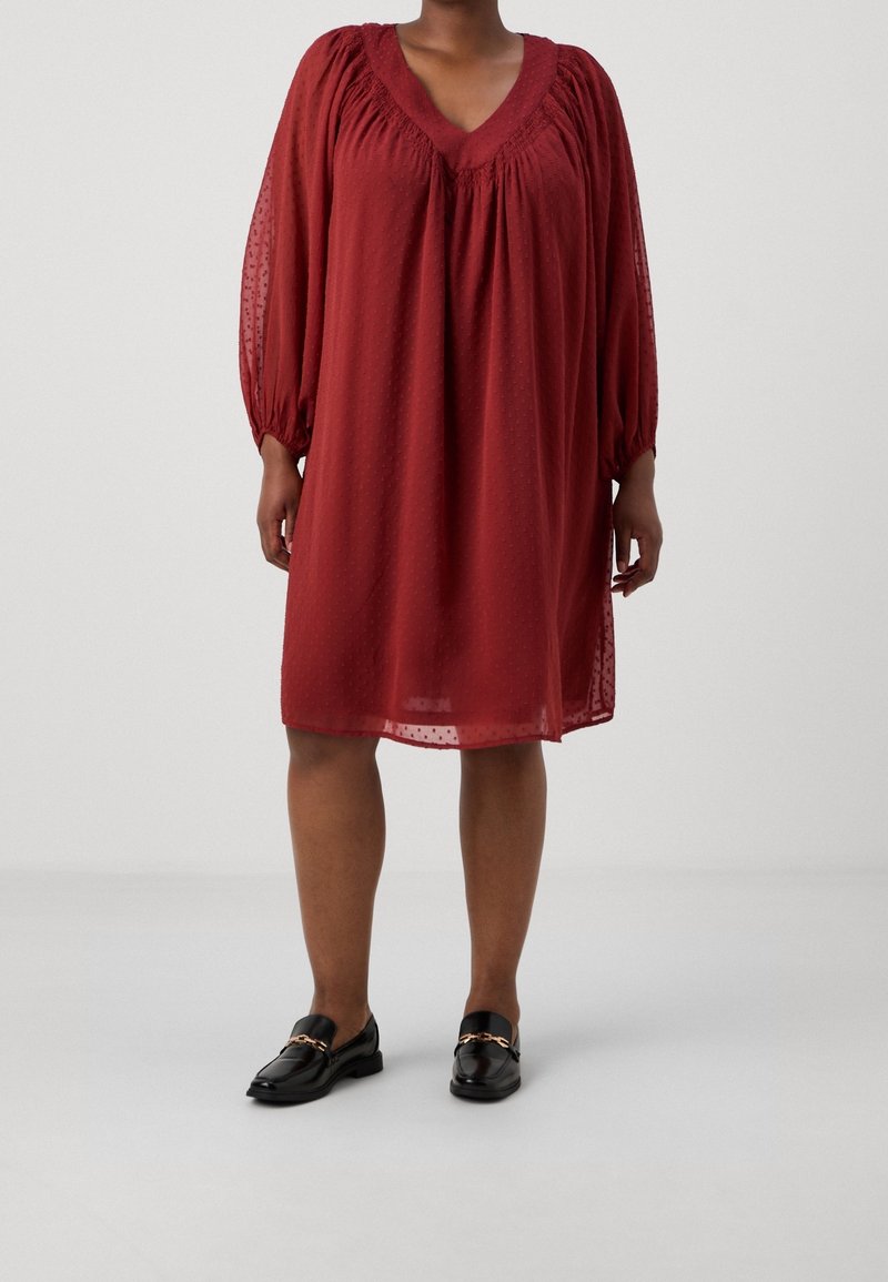 Femme portant une robe ample rouille rouge, longueur jusqu'aux genoux, à manches longues, avec une texture subtile à pois, et des mocassins noirs ornés d'une chaîne dorée.