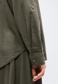 Približen posnetek roke osebe poleg olivno zelene dolgoročne srajce z gumbi na manšetah in usklajenih hlač ali krila iz iste tkanine.